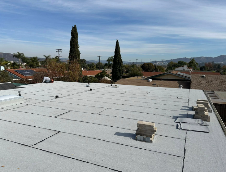 Flat white rooftop with vents, overlooking suburban houses and trees under a clear blue sky.