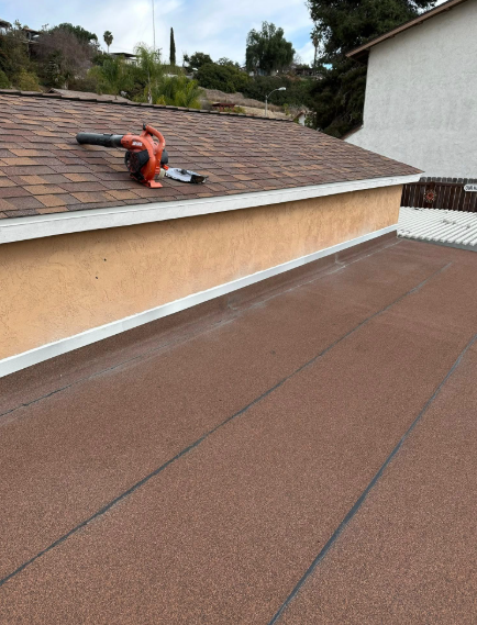 Person using an orange leaf blower on a rooftop with brown shingles and a flat roof edge.
