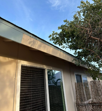 Exterior of a tan house with a sliding glass door, louvered window, and tree branches overhead