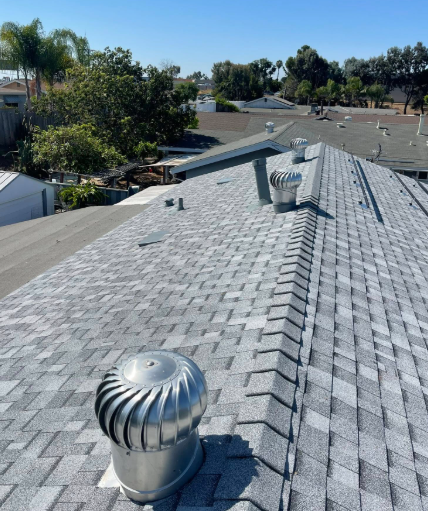 Gray shingled rooftop with silver vents under a clear blue sky, overlooking nearby homes and trees