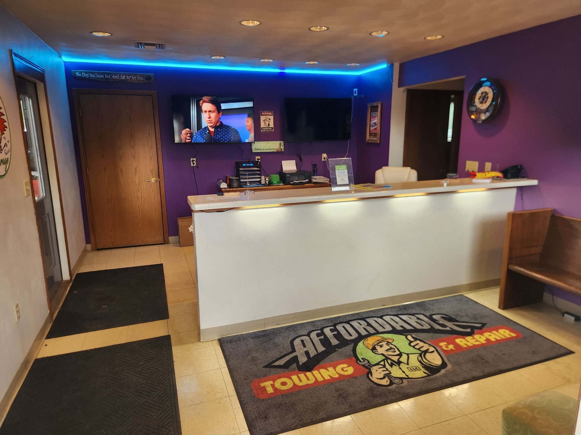 Hotel reception desk with purple walls, blue ceiling lights, and a branded floor mat.