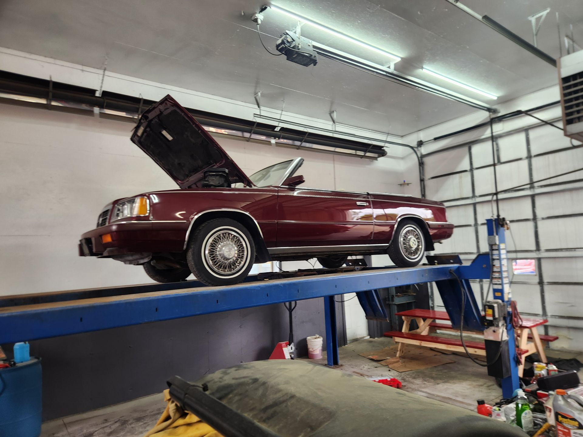 Maroon classic car on a lift in an auto shop, hood open under bright overhead lights