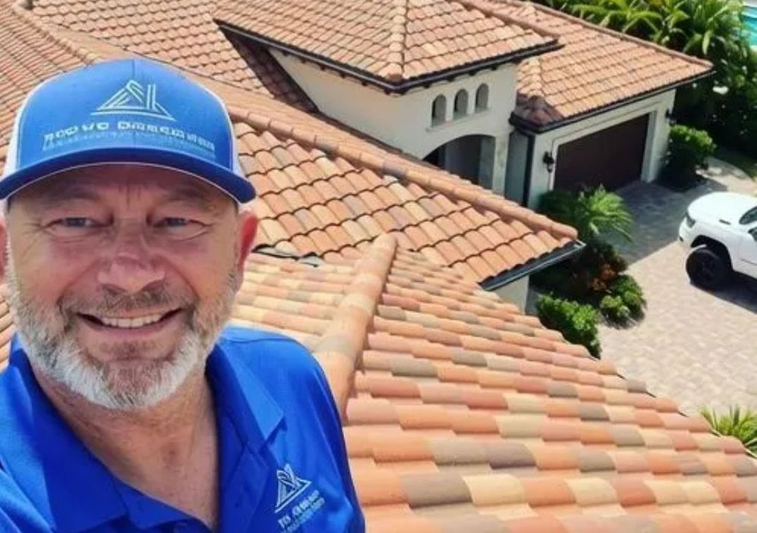 Man on a tiled roof smiles, wearing a company hat and shirt. House and truck in the background.