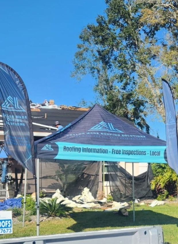 A blue and black tent with roofing company logo is set up in front of a house.