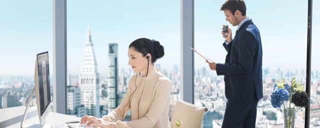 Woman working on a computer with a man standing beside her in a modern office with a city view.