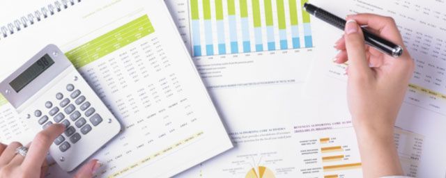 Person calculating on a calculator and pointing at a bar graph. Papers are on a desk.