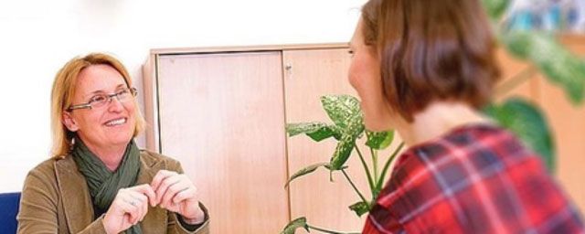 Two women in a room, one smiling while talking. The other is blurred. Plant and cabinets in the background.