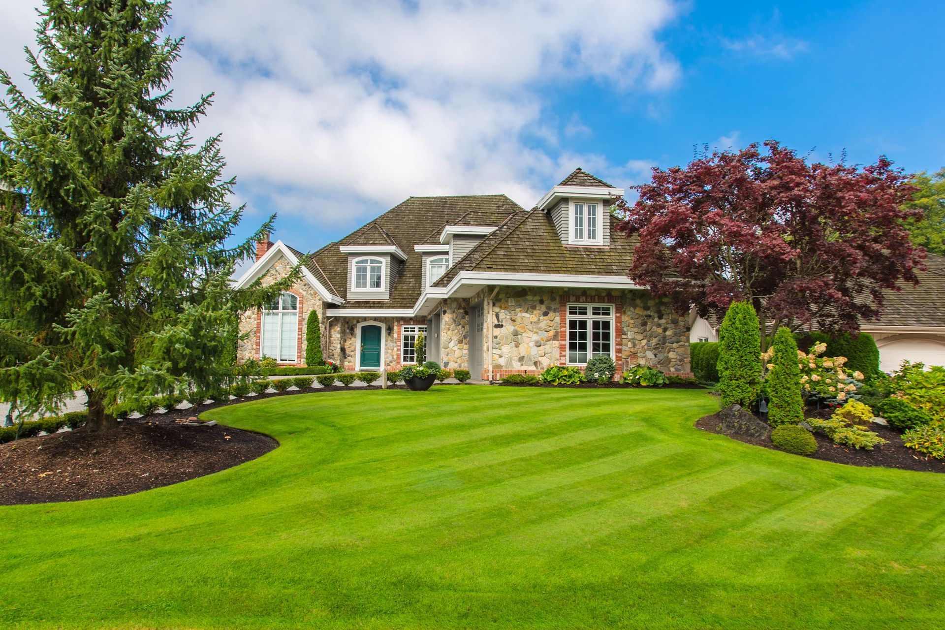 Stone-faced house with manicured lawn and vibrant trees under a partly cloudy sky.