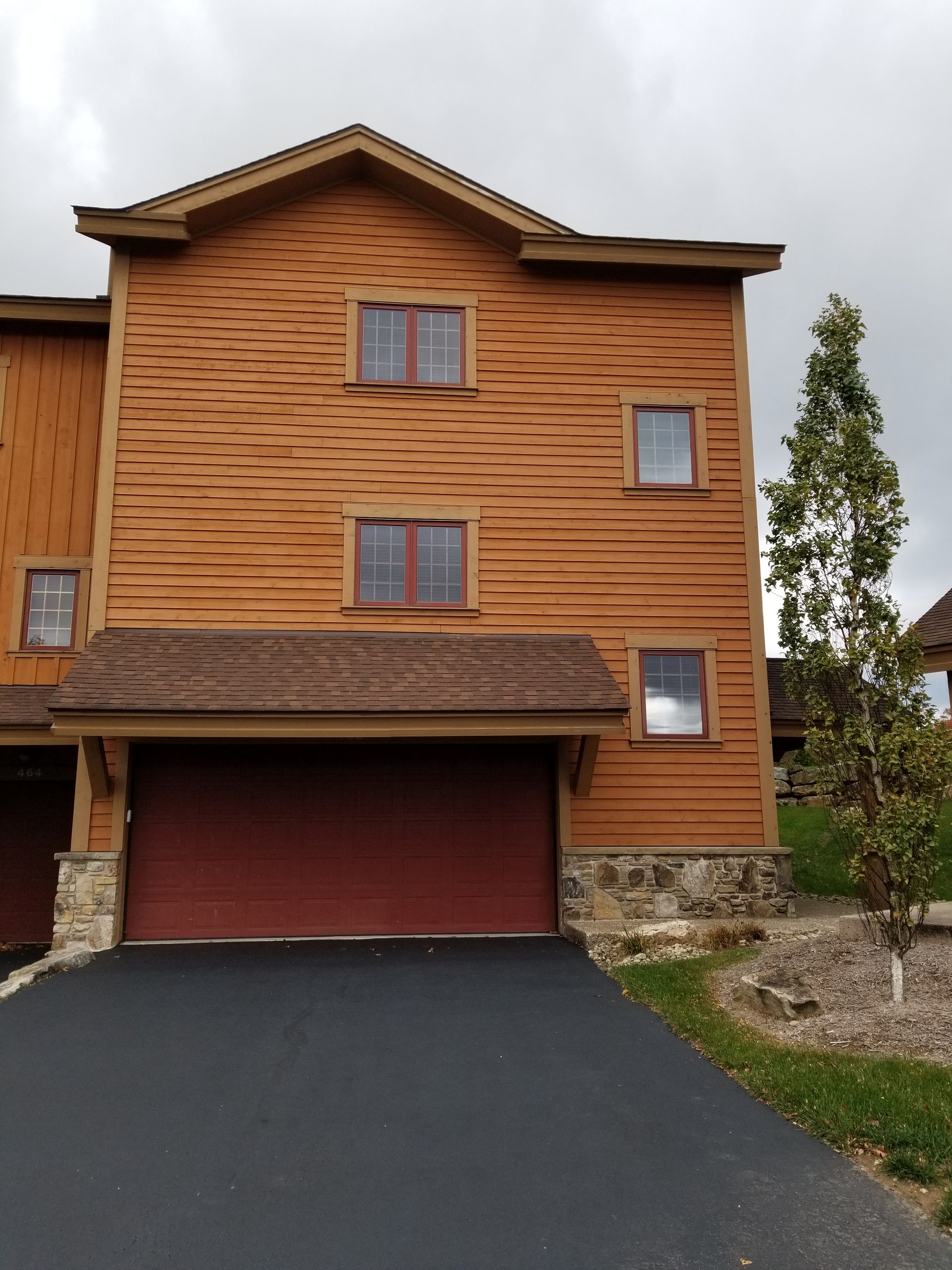 Brown wooden townhouse with a red garage door and windows; driveway, tree.