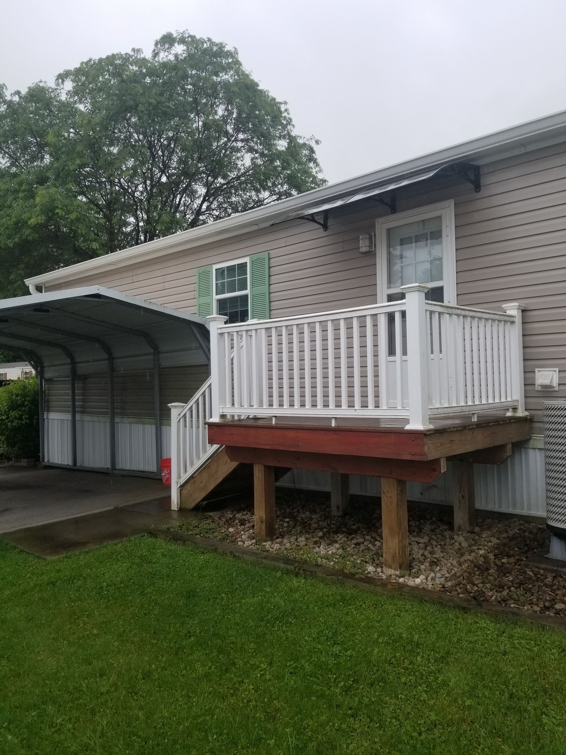 White deck attached to a tan house.  A carport is visible to the left, green grass in front.