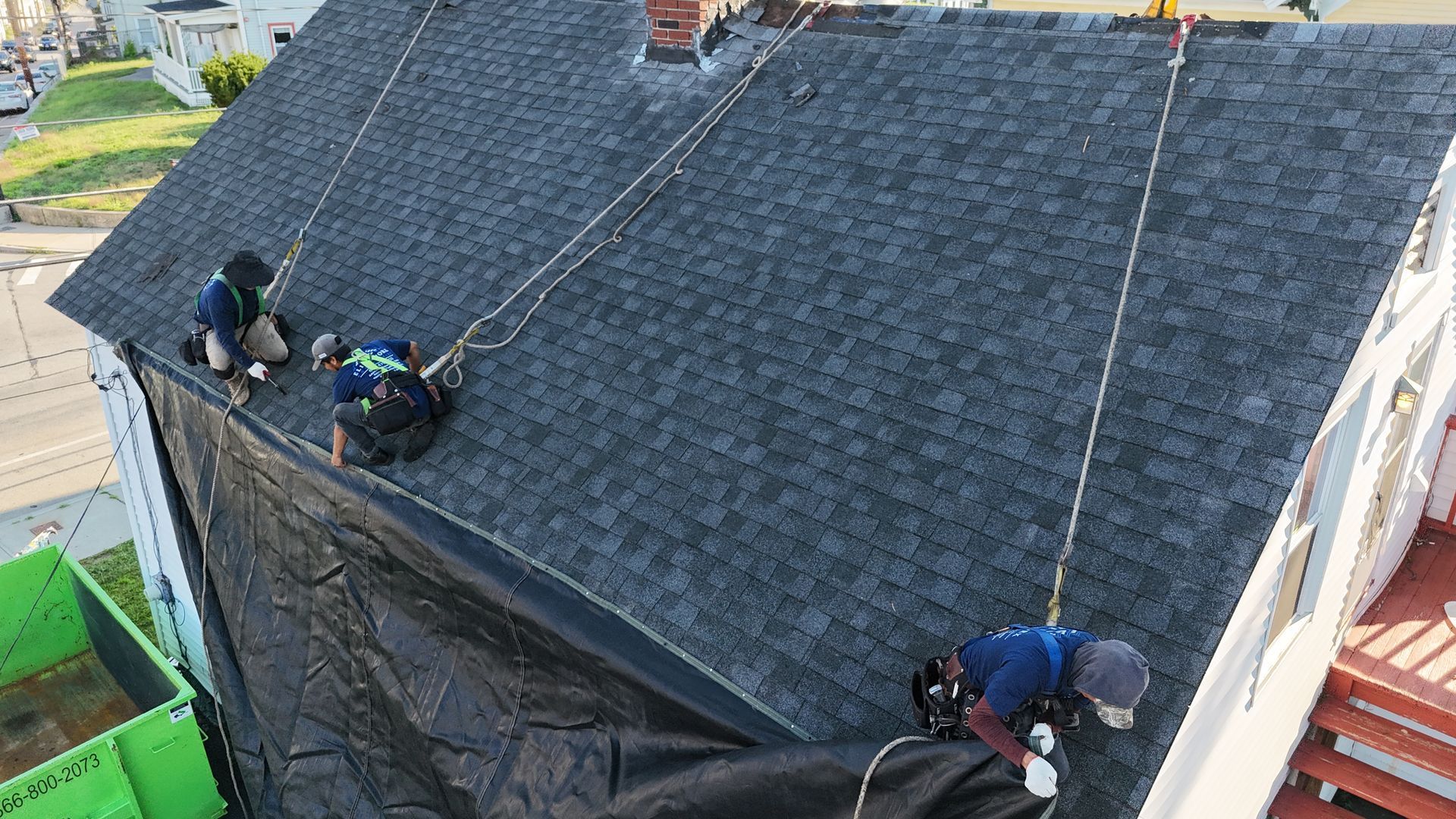 Workers installing black roofing on a steep house roof using safety harnesses