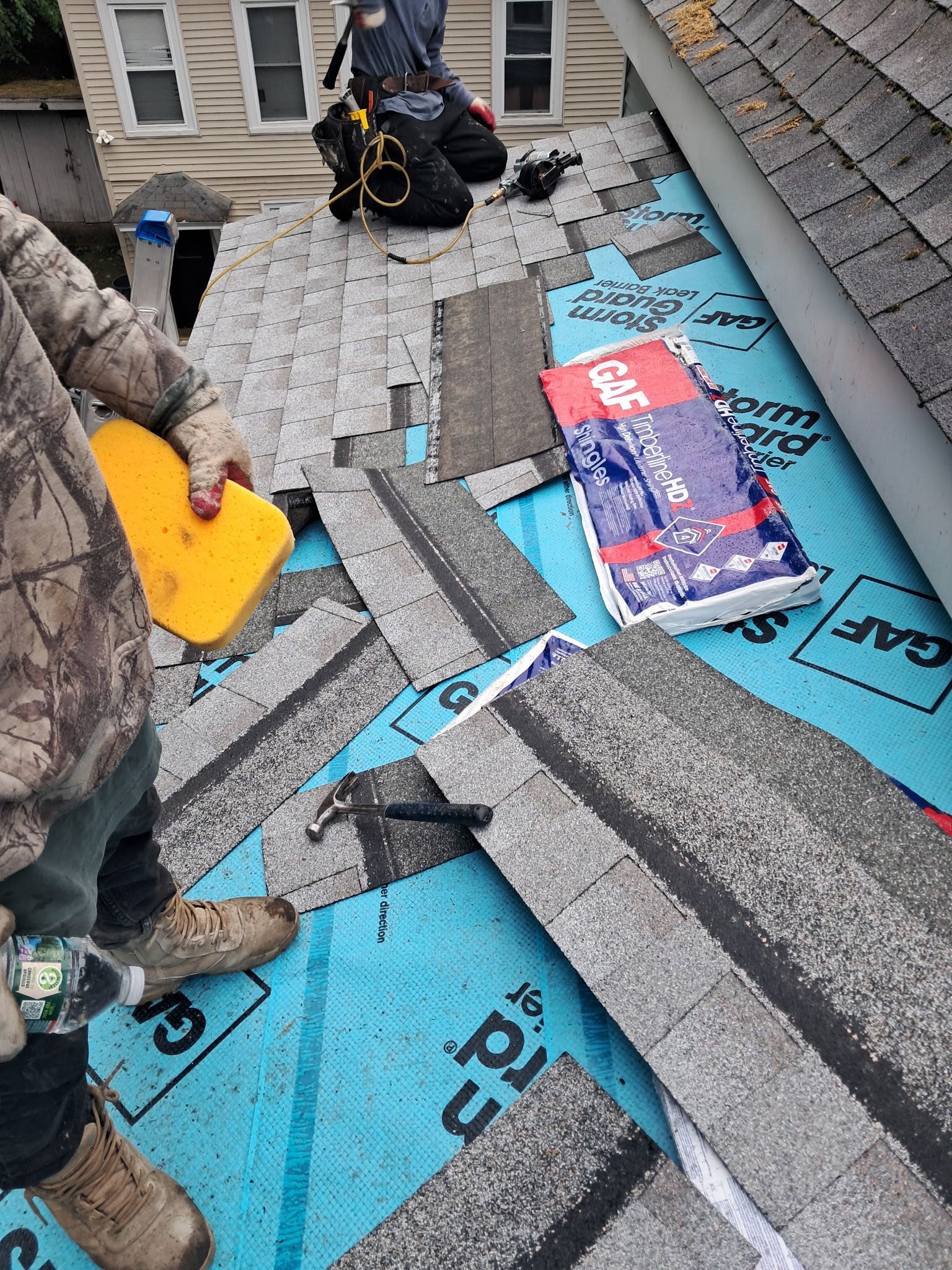 People removing torn blue and gray roof shingles with tools on a sloped rooftop.