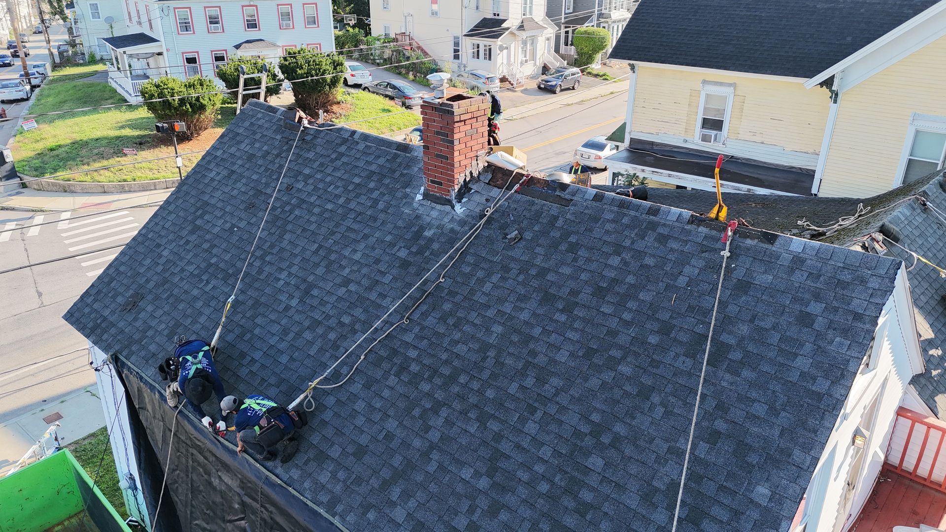 Roofers working on a dark shingled house roof with a brick chimney and safety ropes