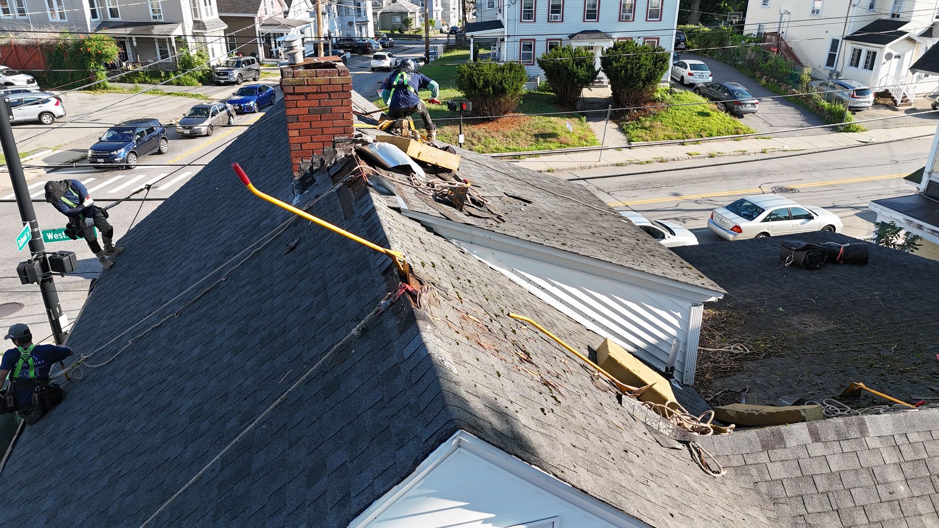 Damaged shingle roof with a chimney and yellow safety line, viewed from above near a street intersection