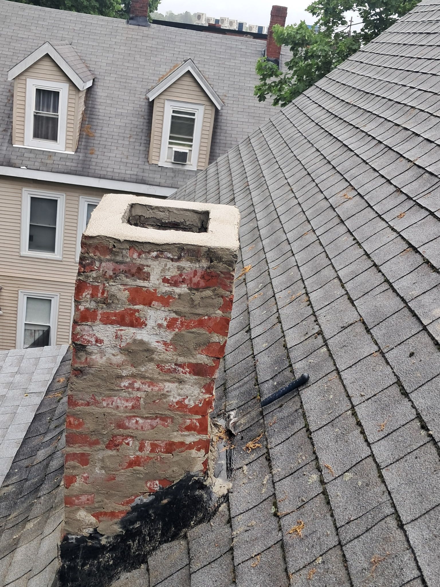 Rooftop with a brick chimney and dormer windows, showing weathered shingles and black roof tar repair.