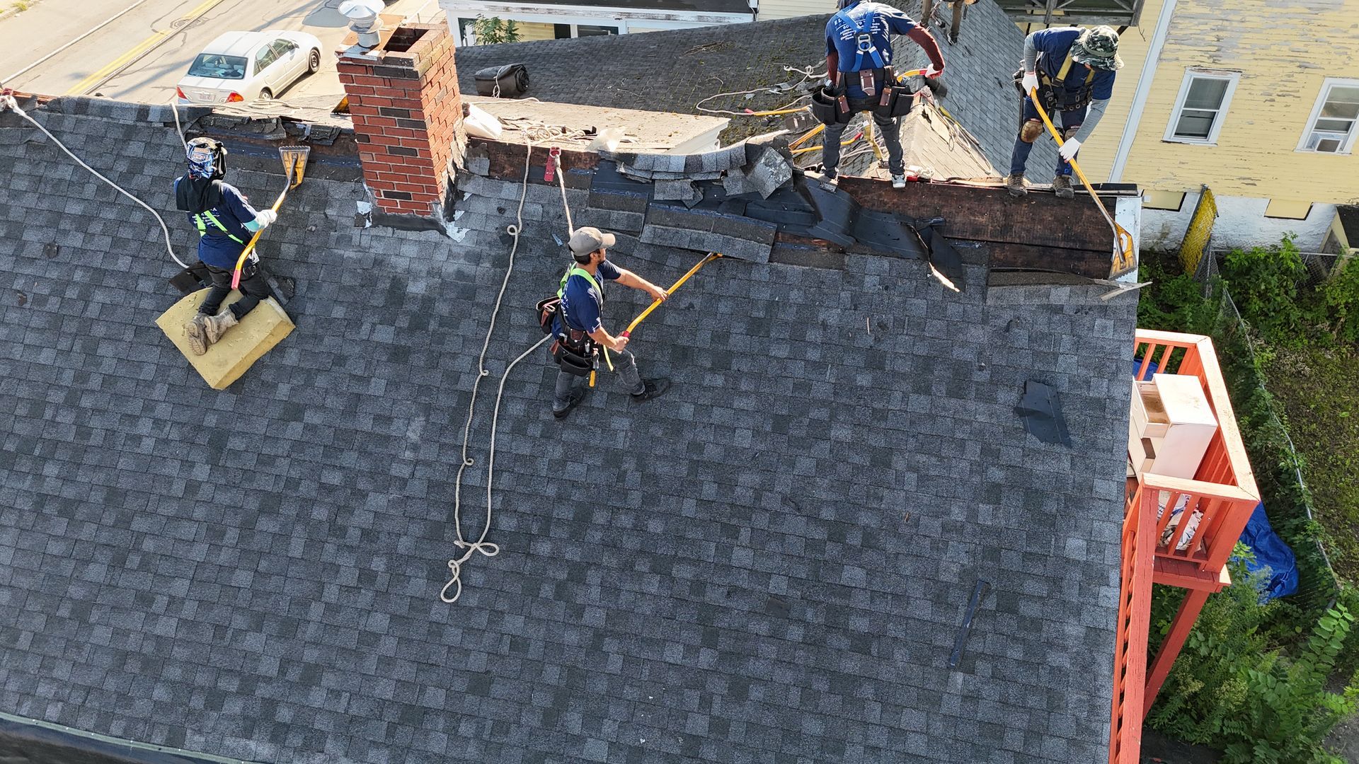 Roofers repairing a dark shingle roof near a brick chimney, with ropes and tools visible