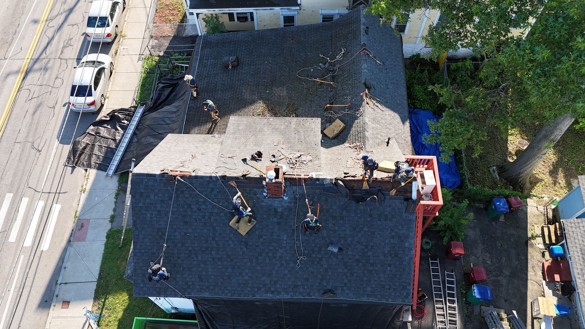 Aerial view of a flat-roof building with workers and equipment, beside a street and trees
