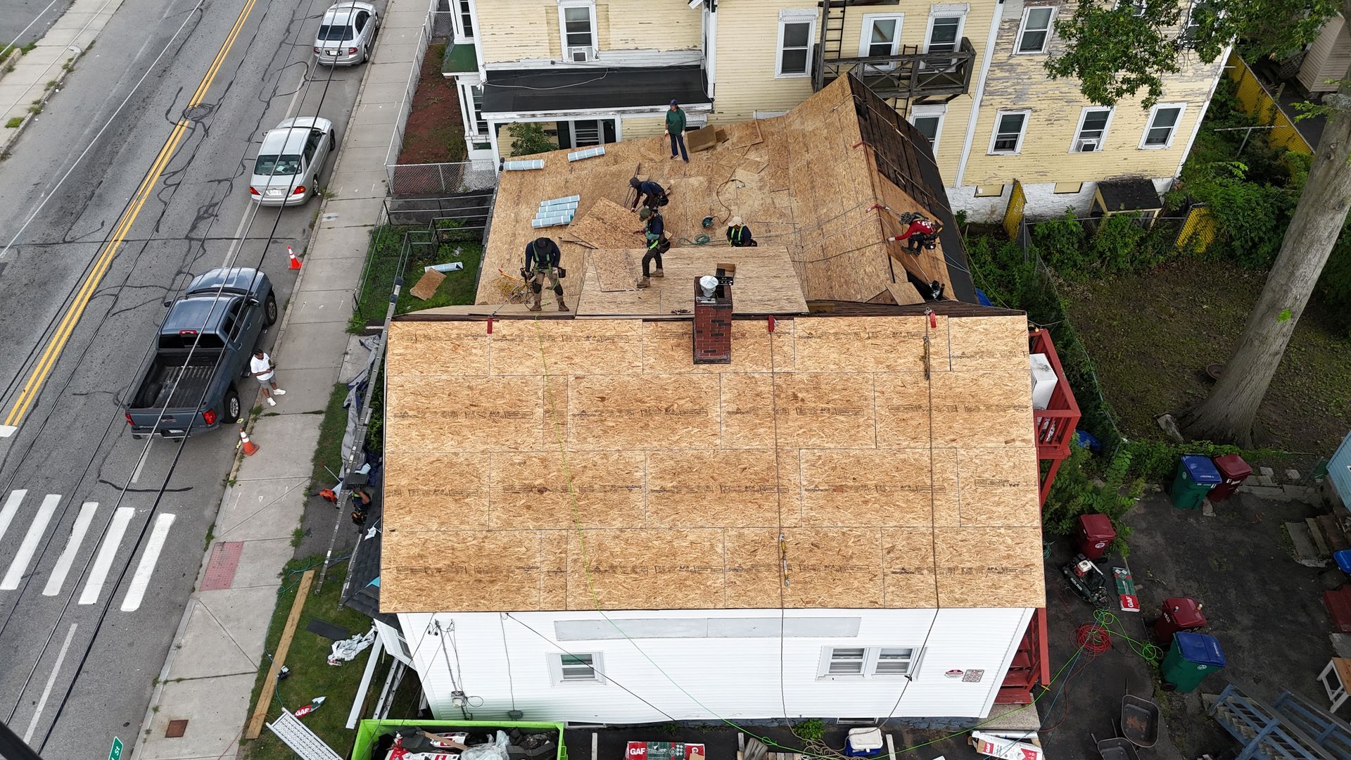 Roofers working on a tan-roofed house beside a street with parked cars and sidewalks
