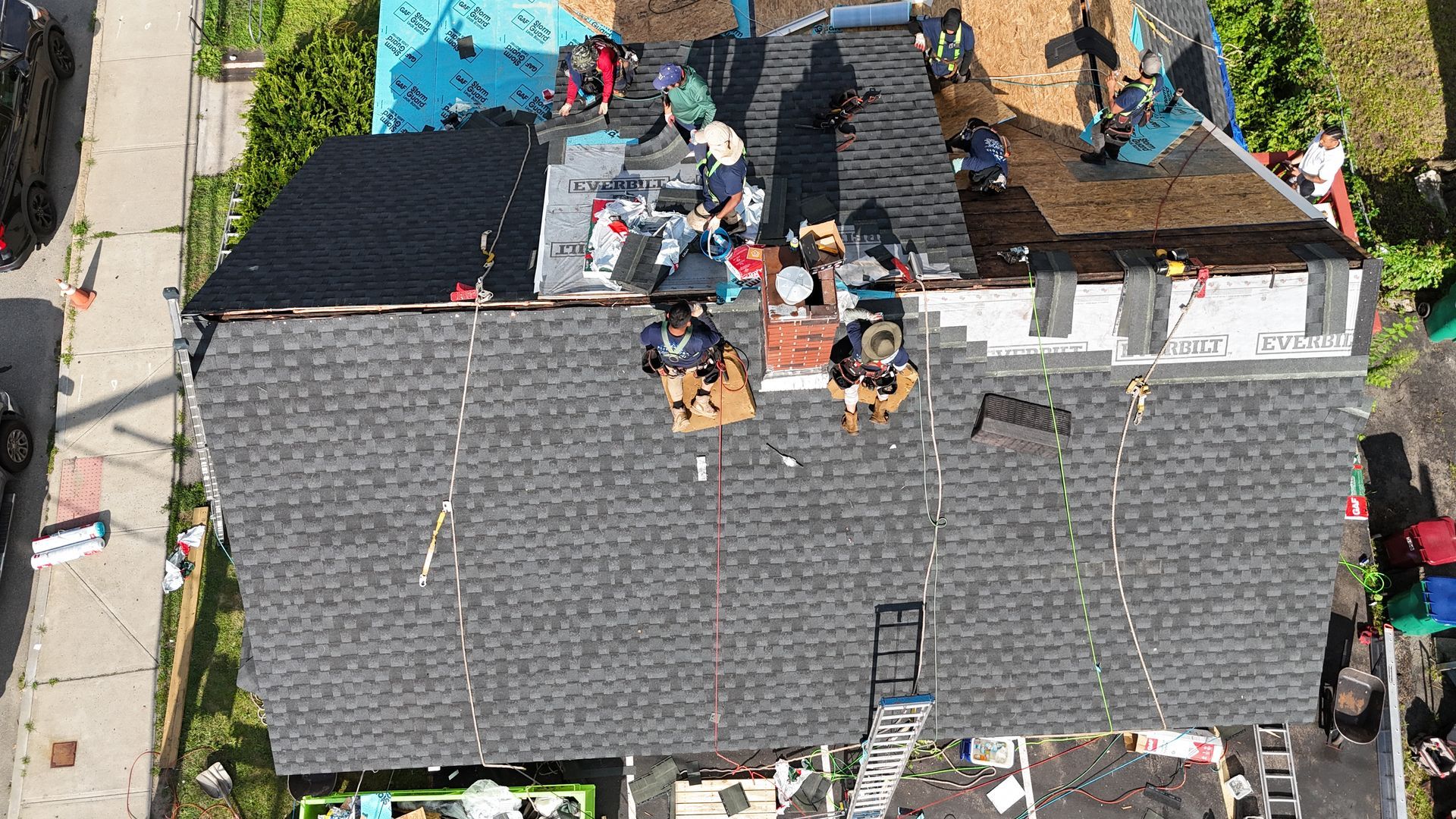 Aerial view of people working on a shingled rooftop with tools and materials nearby
