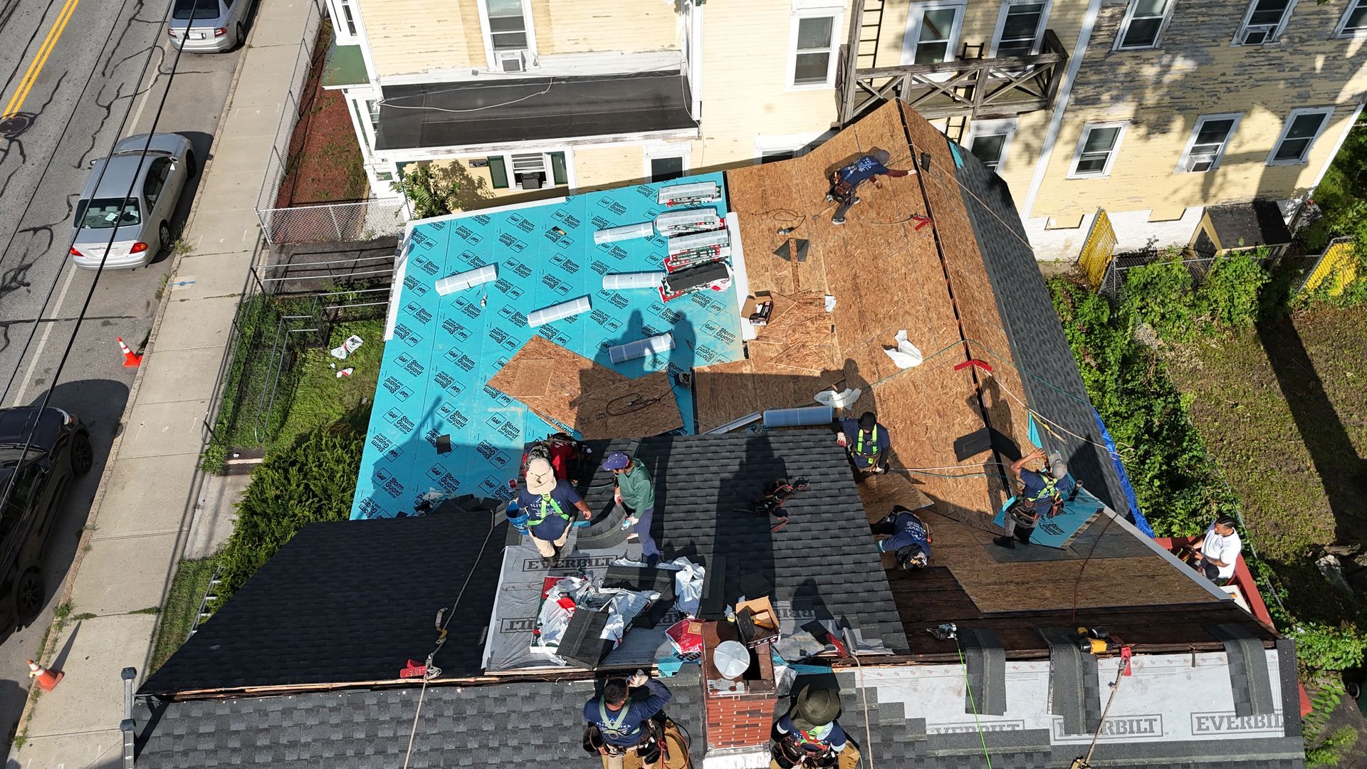Rooftop workers installing blue tarps and black roofing panels on a damaged building roof