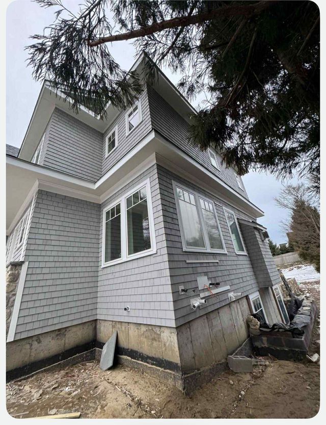 Gray house exterior with white trim, viewed from below, with evergreen branches overhead and a basement foundation visible