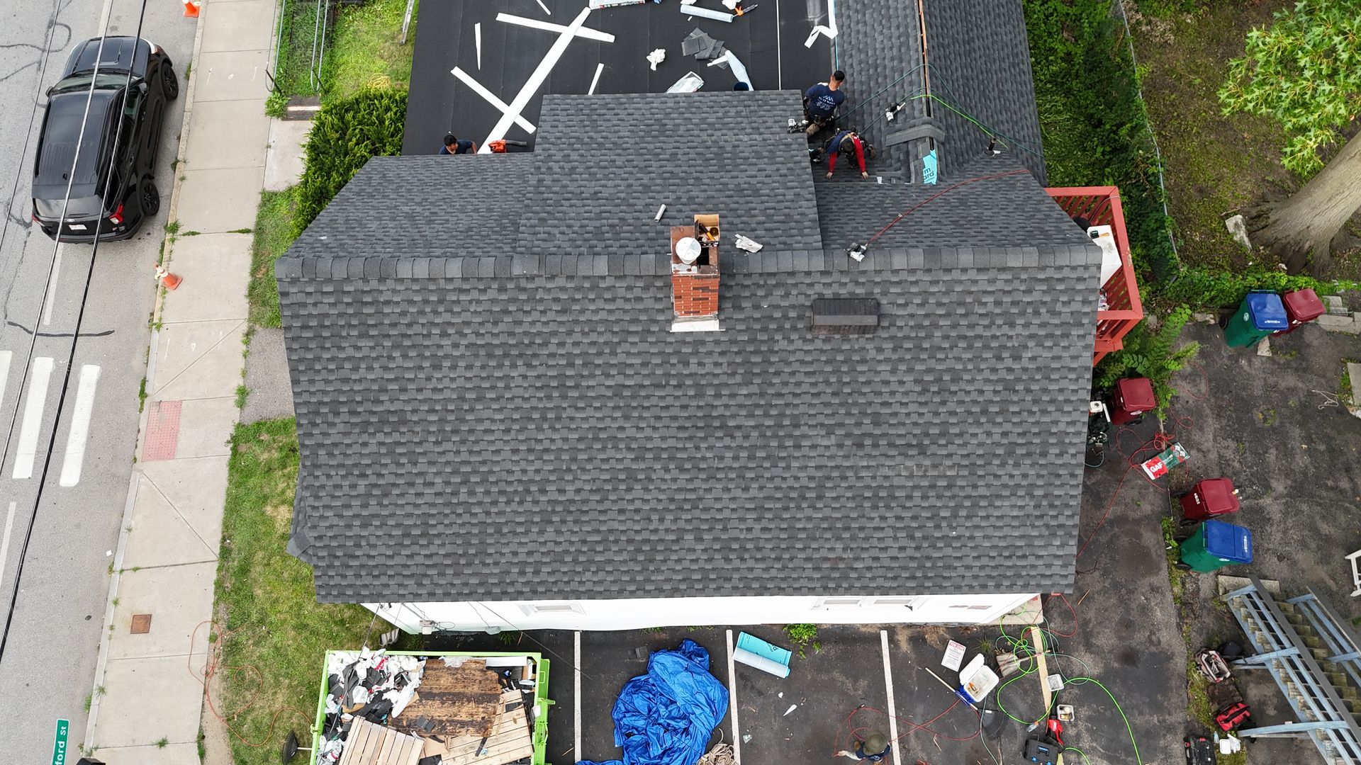 Aerial view of a house roof beside a street, with a driveway, sidewalk, and scattered items in the yard