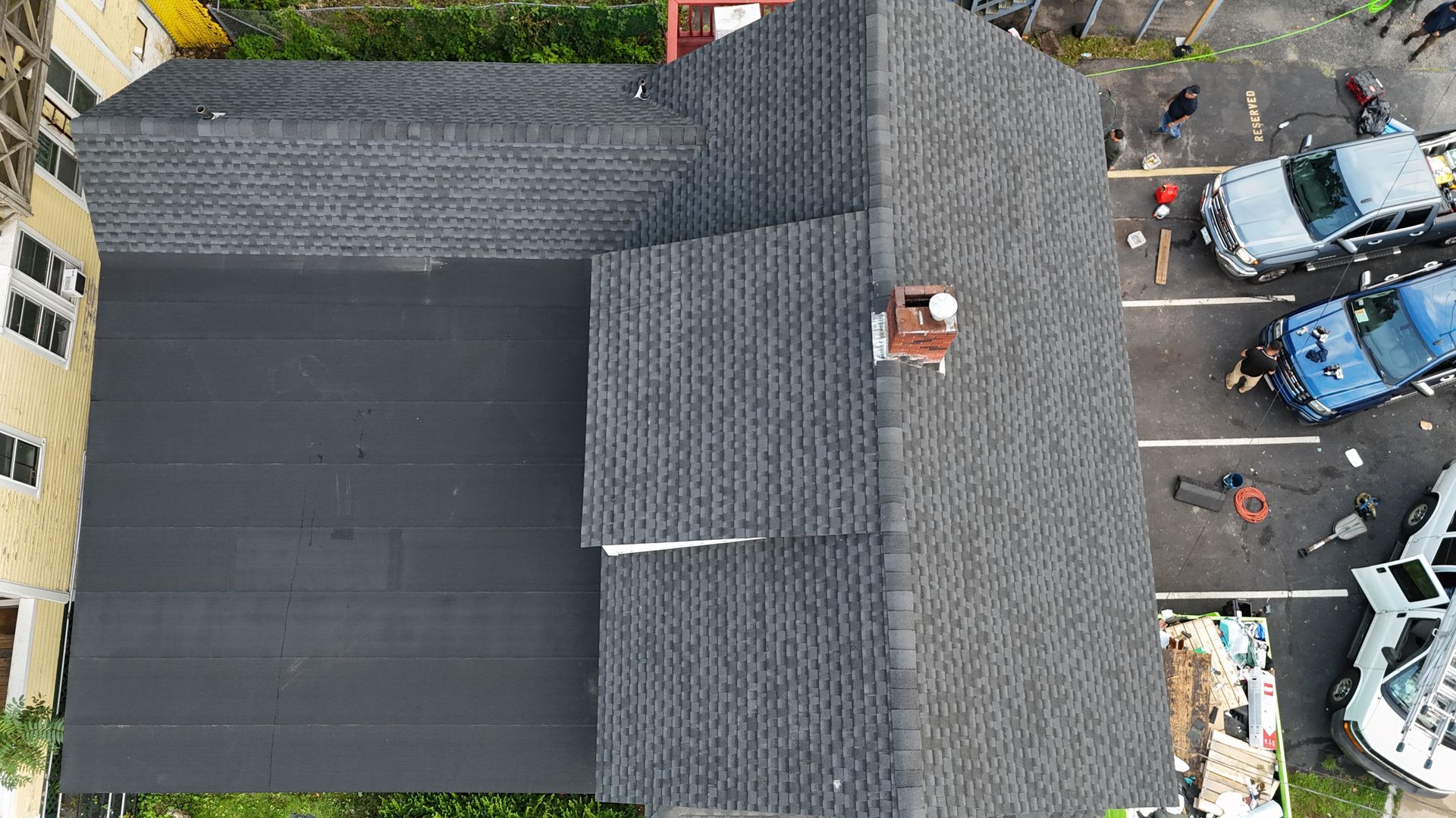 Aerial view of a house roof with dark shingles and a paved parking lot with cars beside it