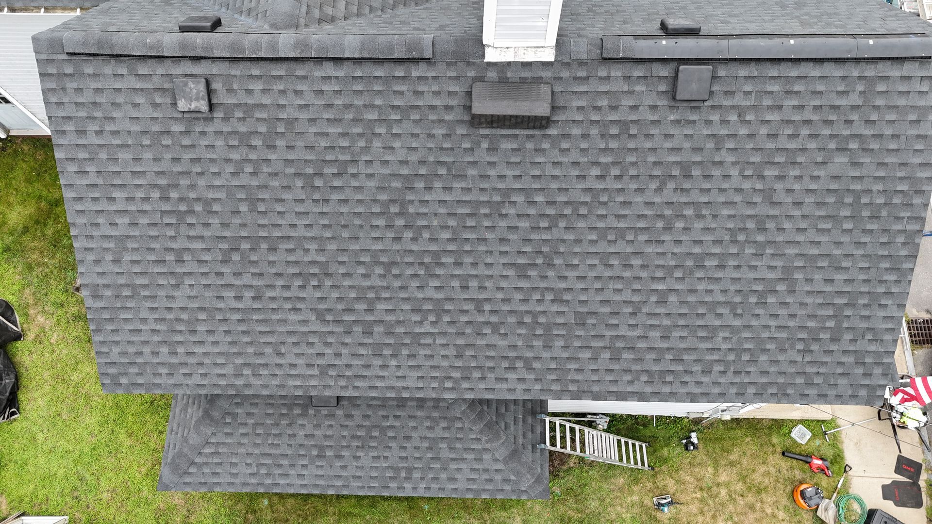 Aerial view of a gray-shingled house roof with a small ground-level patio and yard visible below