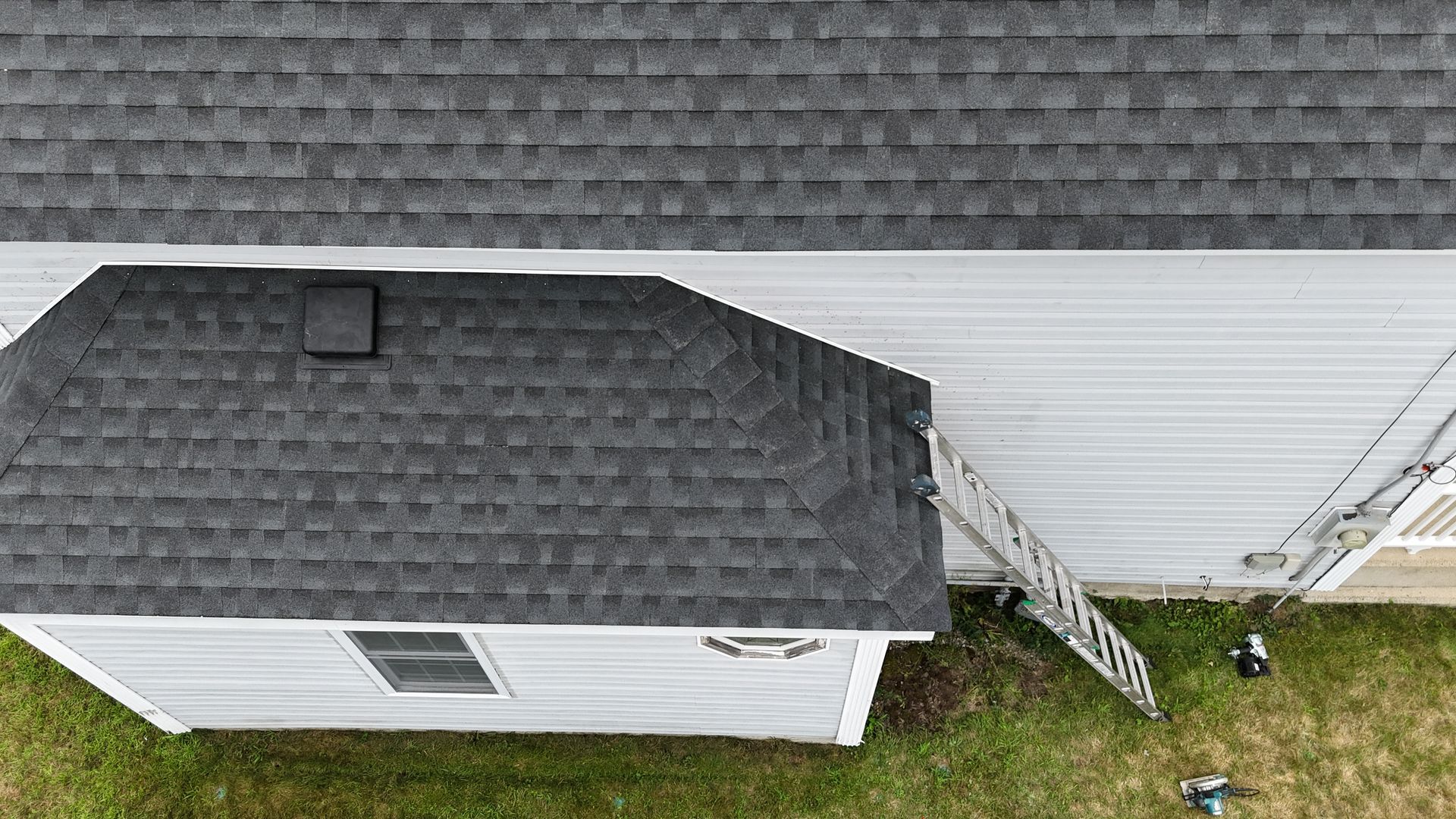 Aerial view of a gray-shingled house roof with a white side wall, ladder, and grassy yard below
