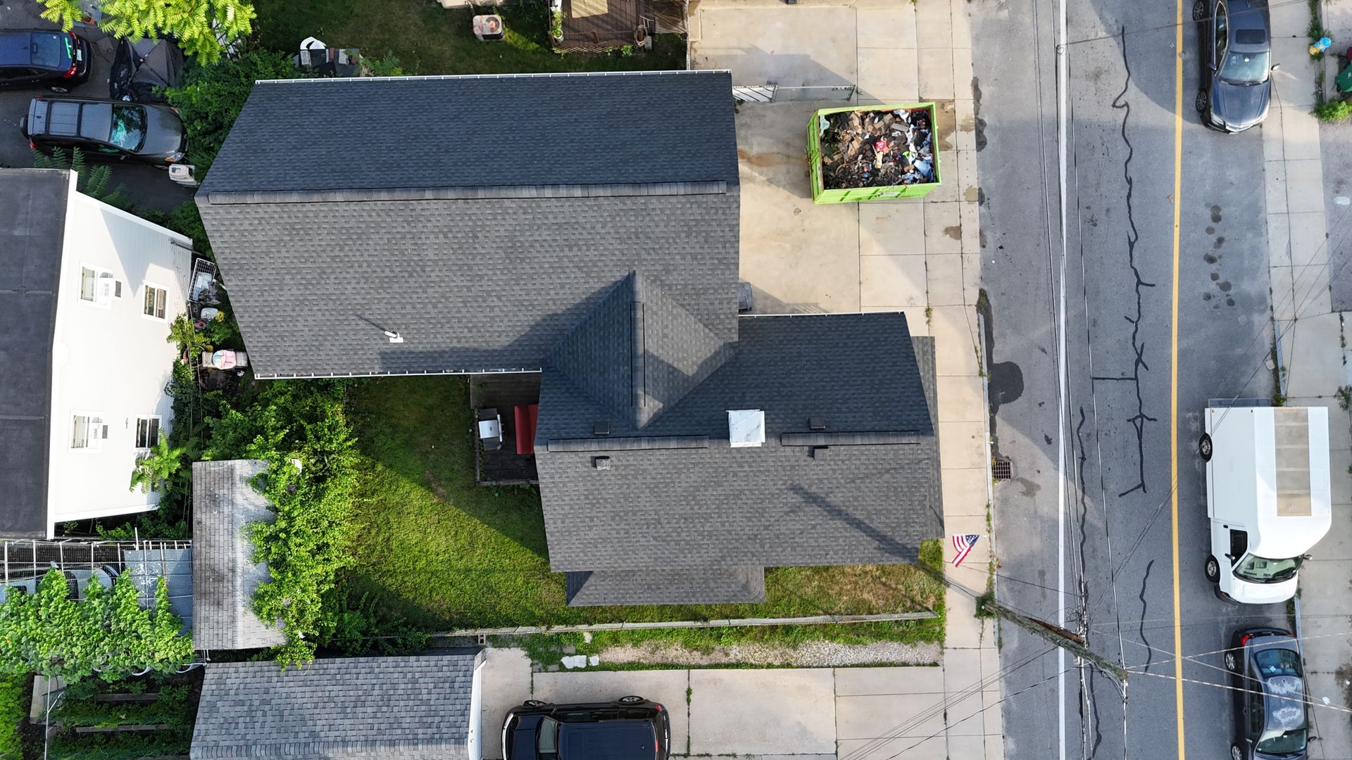 Aerial view of a house with gray roof, driveway, yard, and parked vehicles along a street