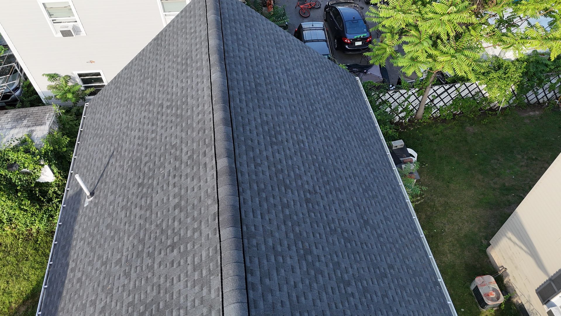 Aerial view of a gray-shingled roof beside a yard, with a car and trees nearby