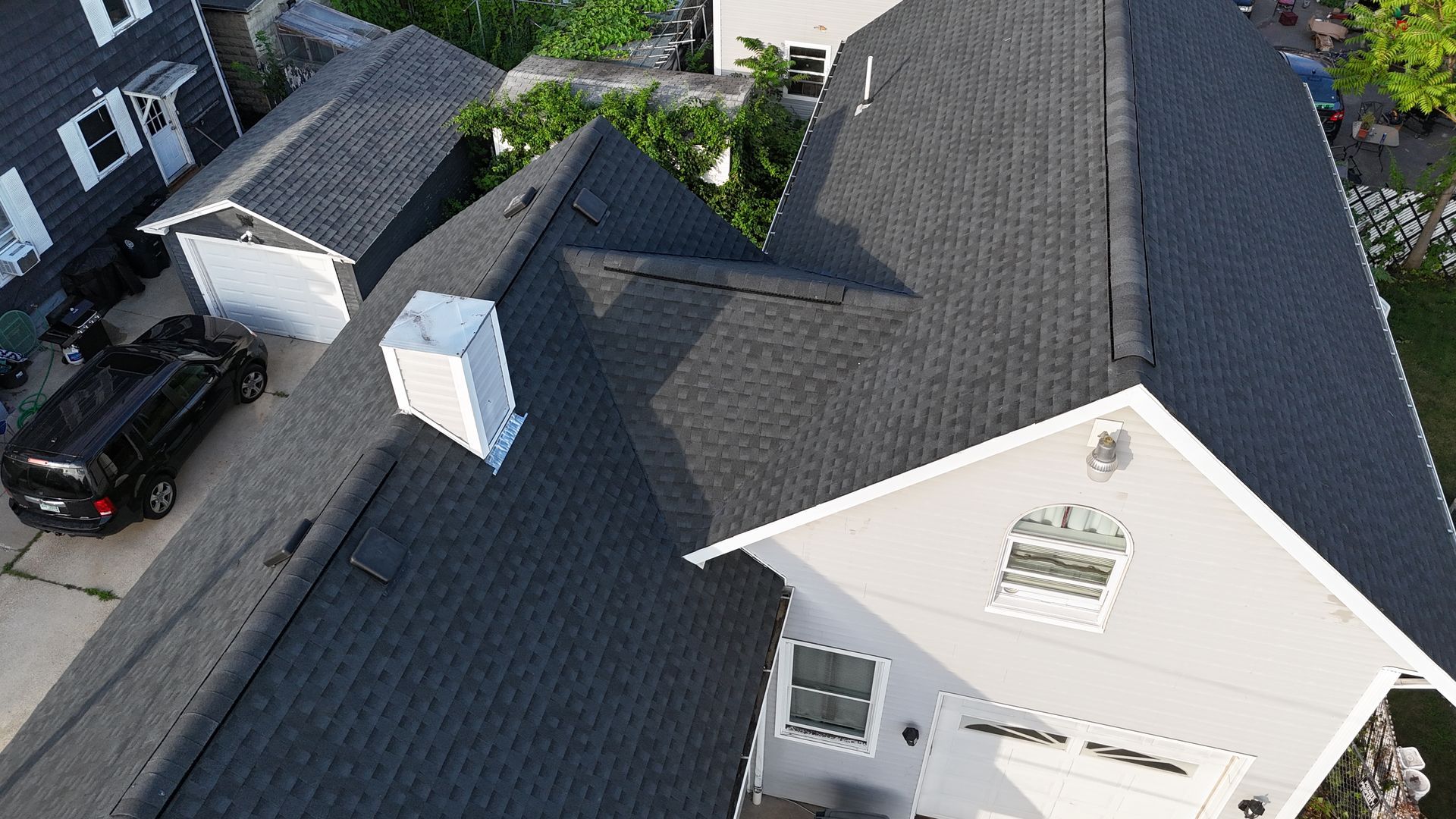 Aerial view of a house with dark gray roofs and a white gable, beside neighboring homes and a driveway