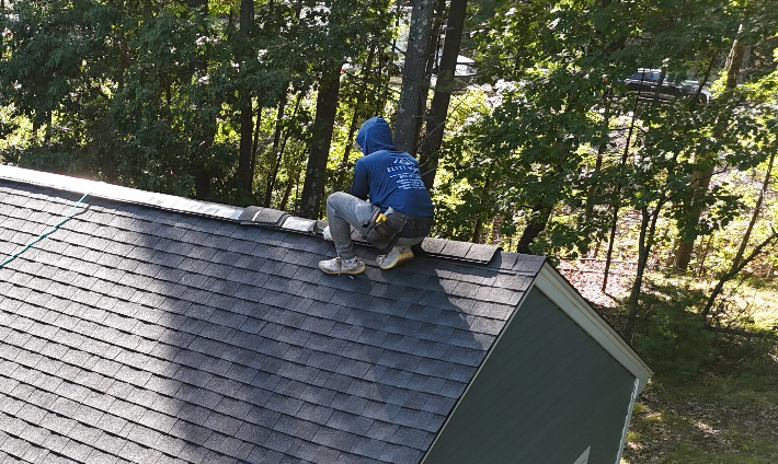 Person in blue jacket crouches on a gray shingled roof beside trees.