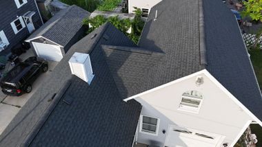 Aerial view of a gray-shingled house roof with dormer windows and nearby parked cars