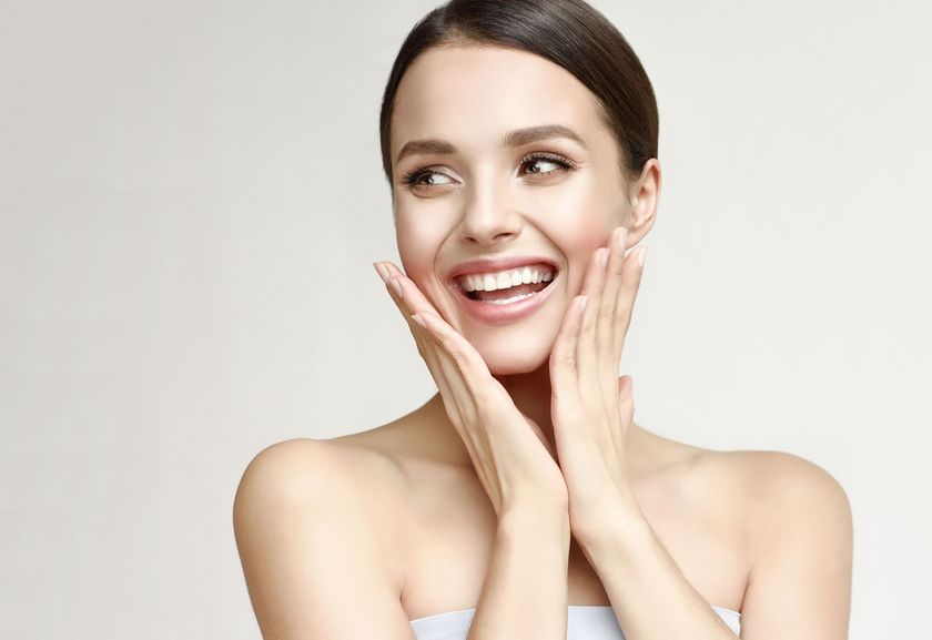 A woman with fair skin smiles widely, touching her cheeks, against a light background.