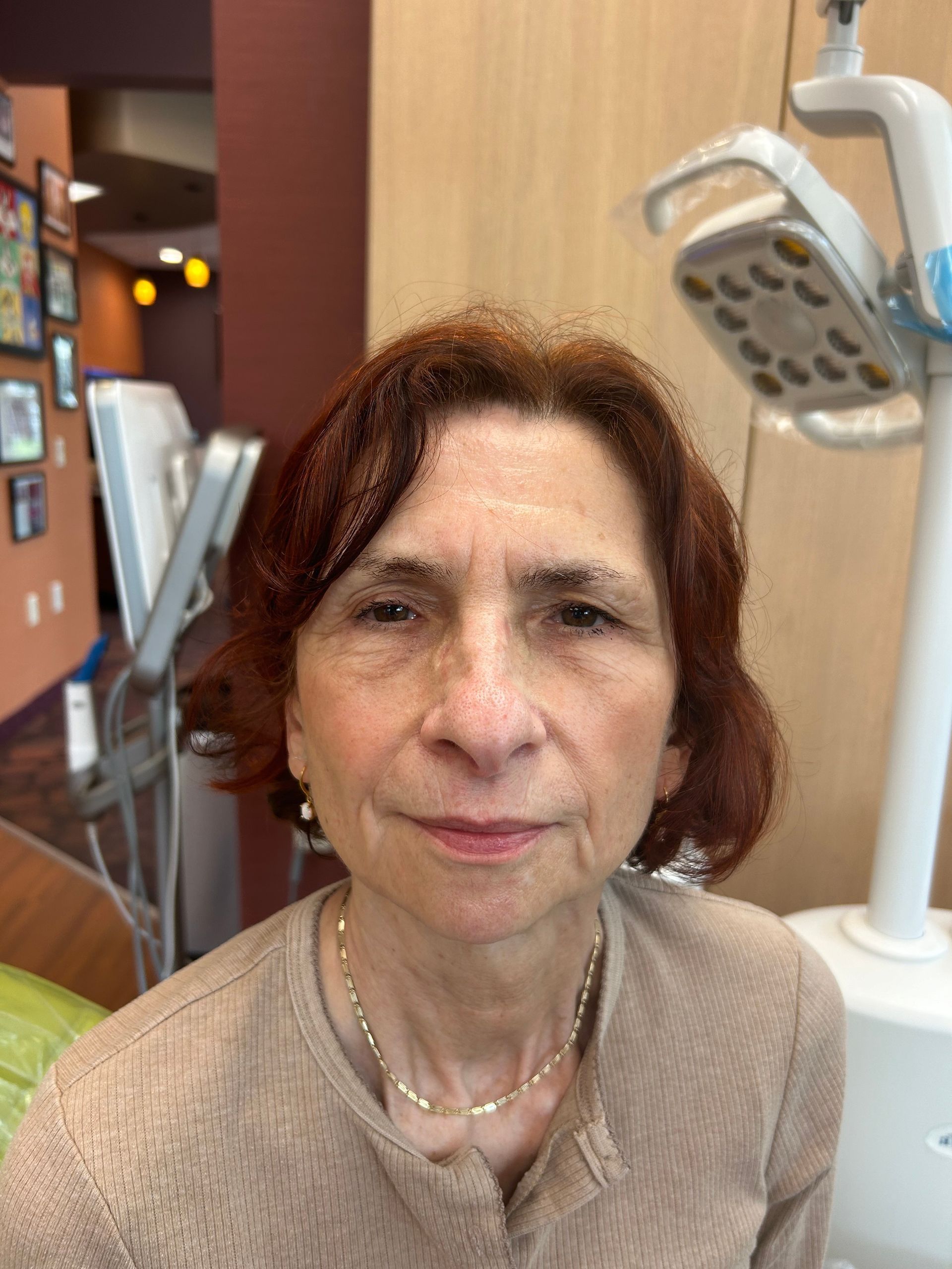 Woman in beige sweater at a dental office, posing for the camera, dental equipment visible.
