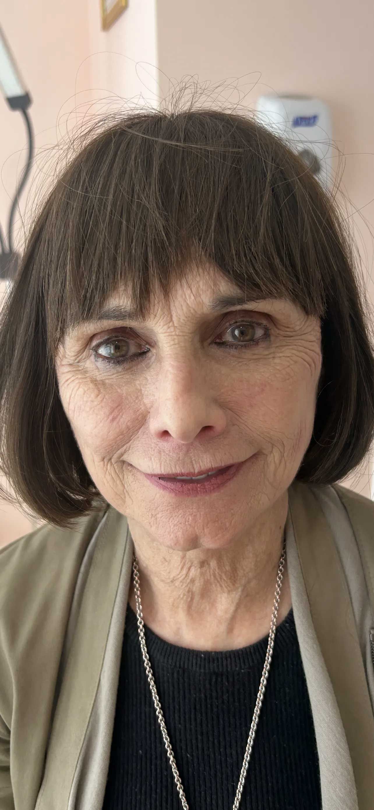 A person with short brown hair, wearing a gray sweater and a necklace, looking at the camera.