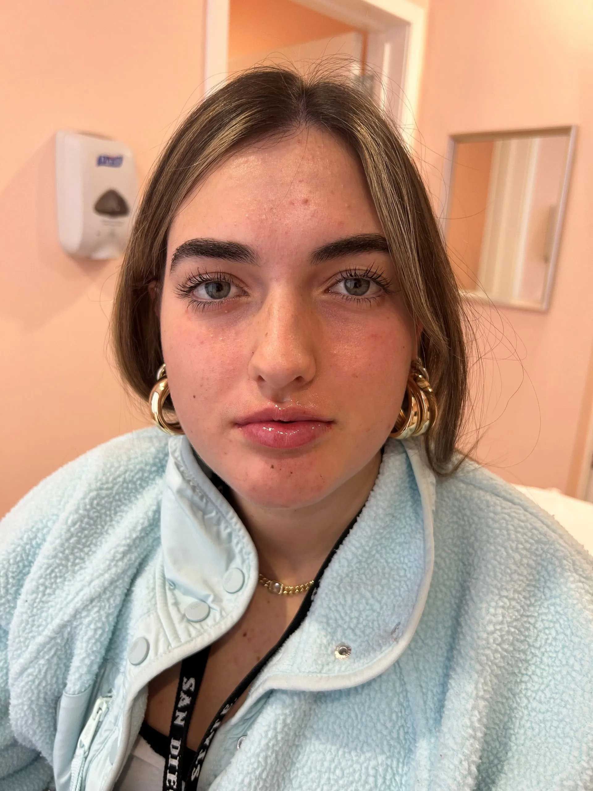 Woman with light skin, blue eyes, wearing gold earrings, light blue fleece, and a small necklace, indoors.