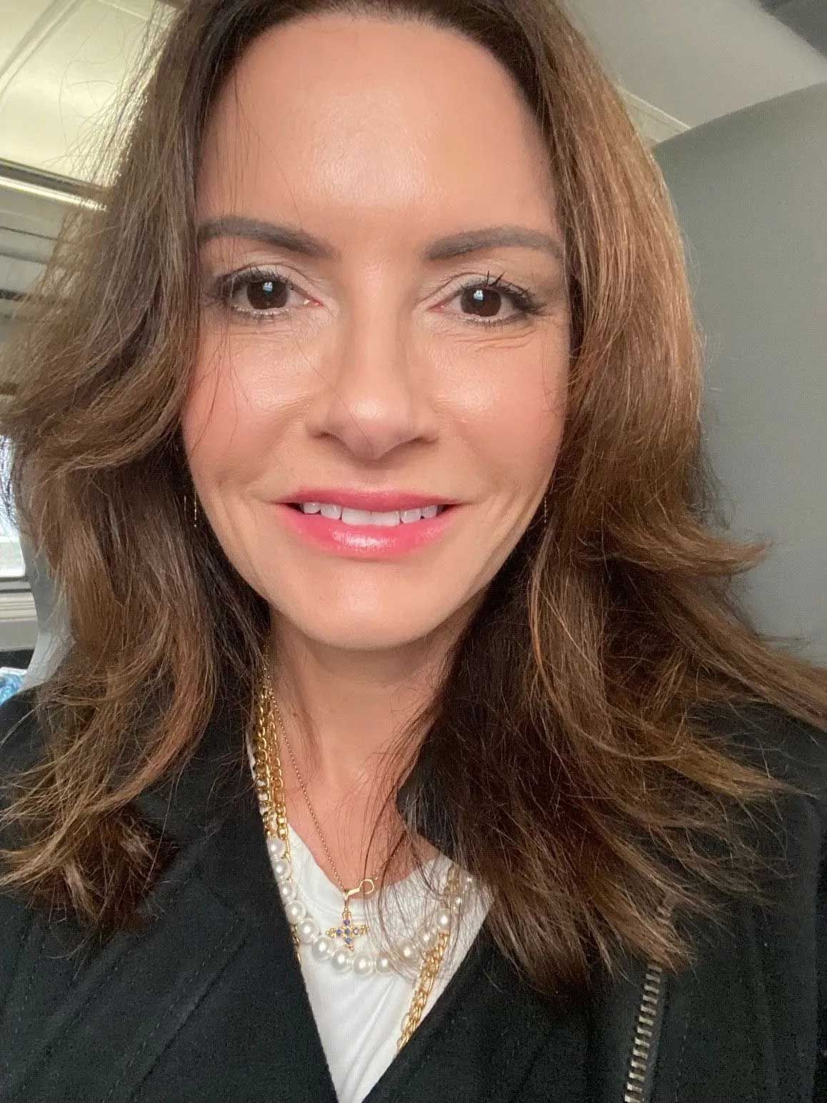 Woman with brown hair and pink lipstick smiles, wearing a white top, black blazer, and gold necklace.