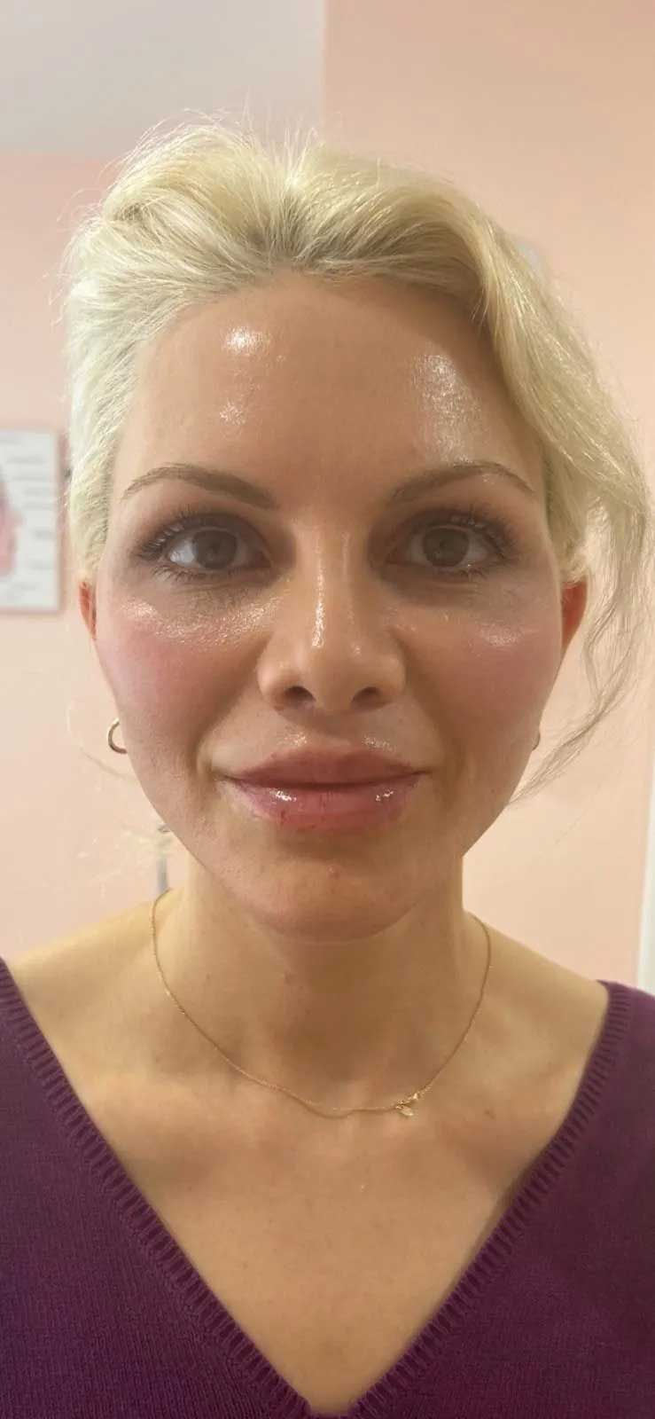 Blonde woman with fair skin, pink blush, and a gold necklace smiles at the camera in a bright room.