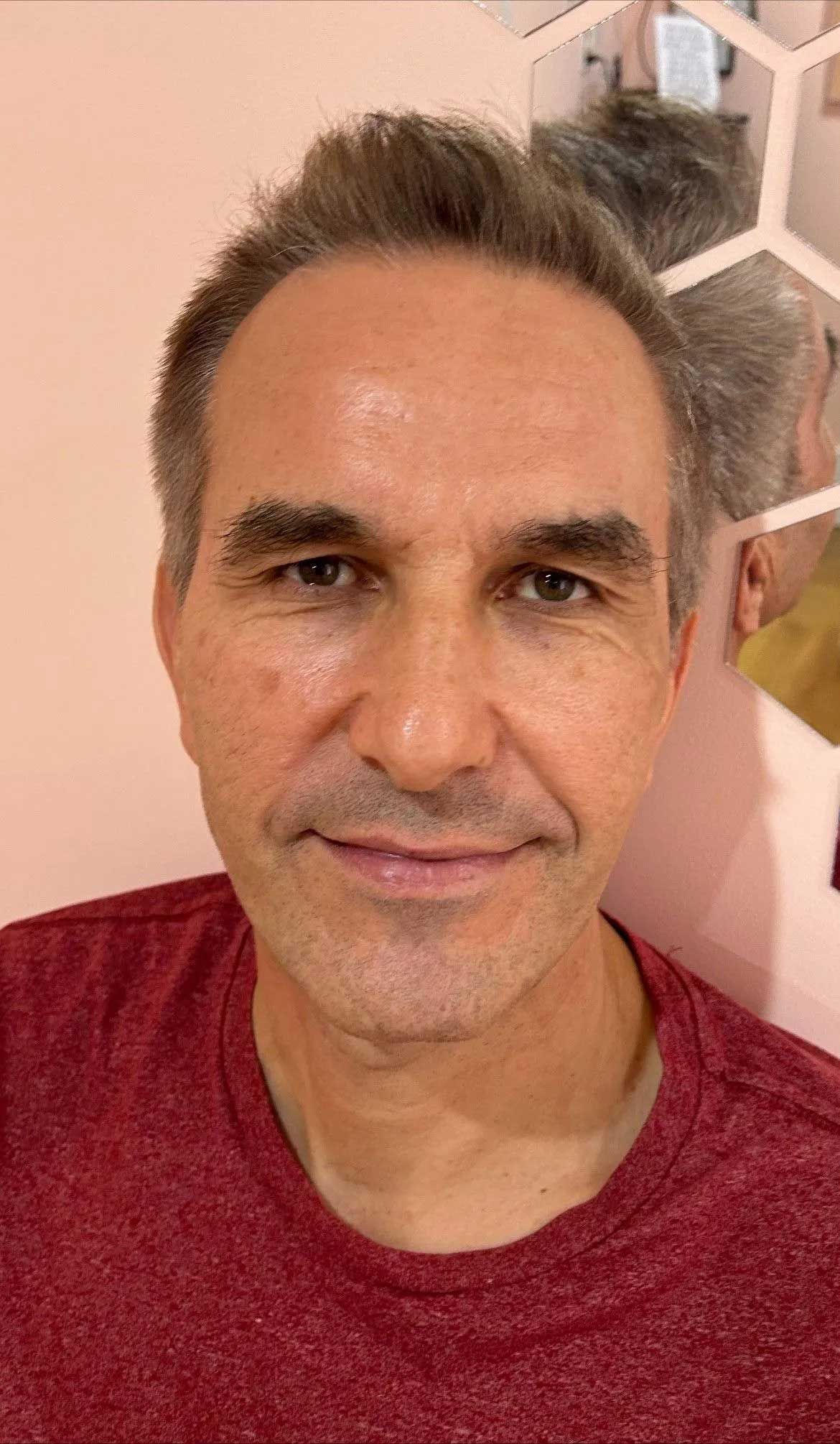 Middle-aged man with graying hair smiles at the camera in a pink room. He wears a red shirt.