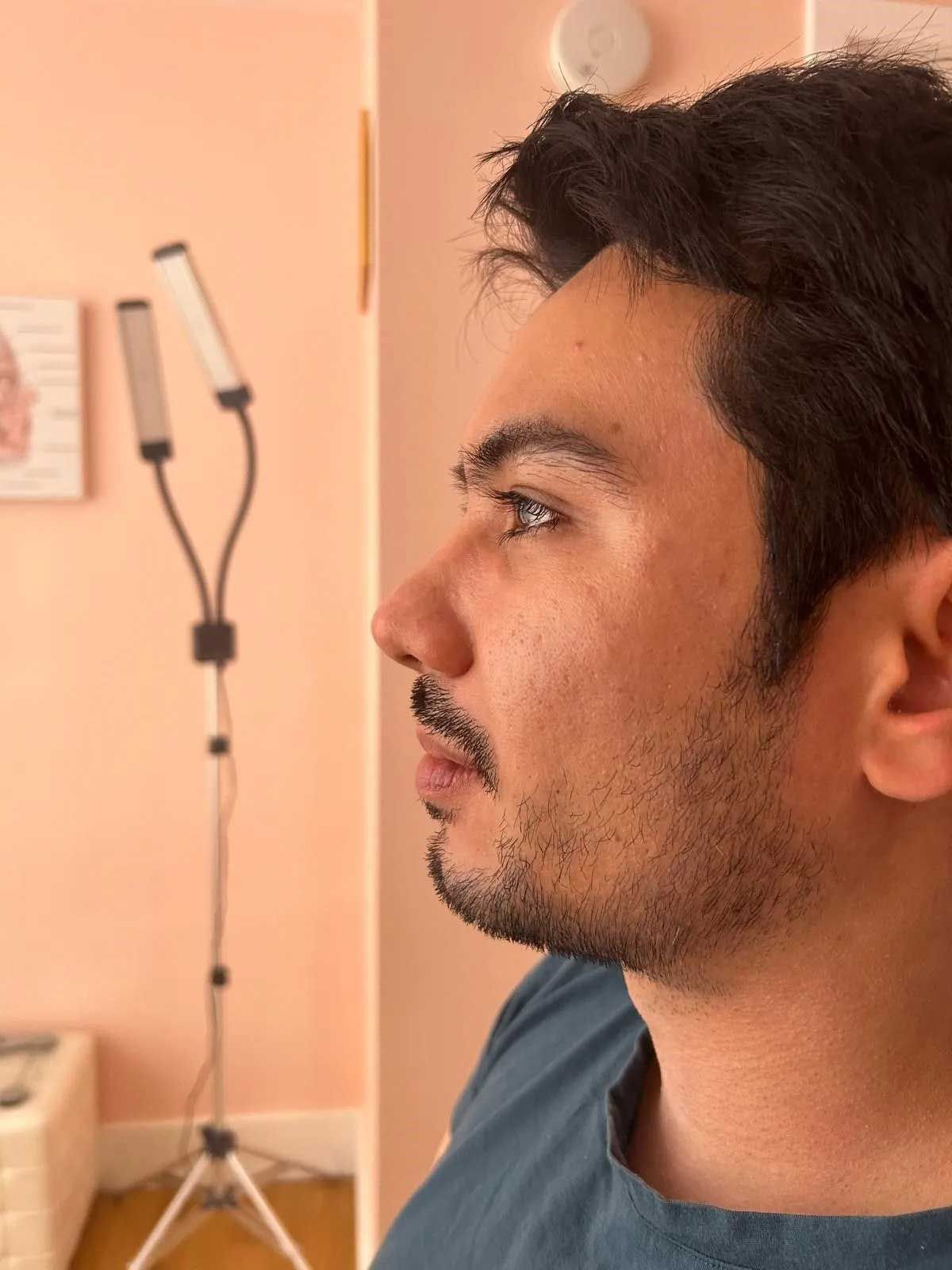 Man with dark hair and beard in profile. He is in a pink room with a lamp.