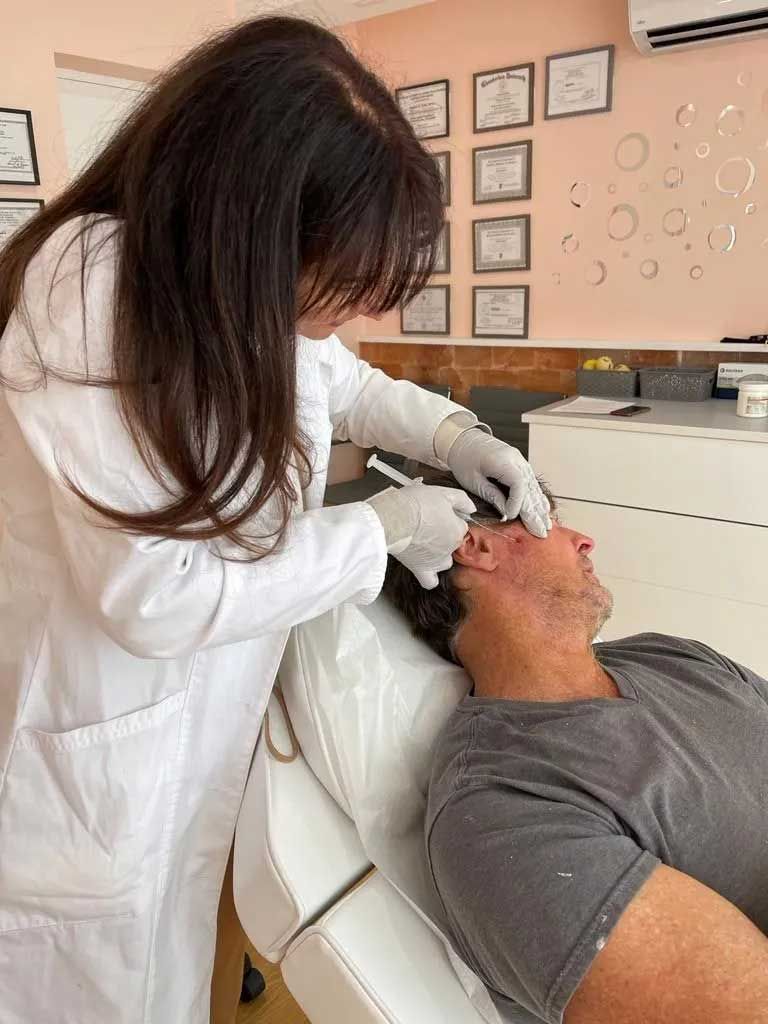 A doctor in a white coat injects a syringe into a man's face in a medical setting.