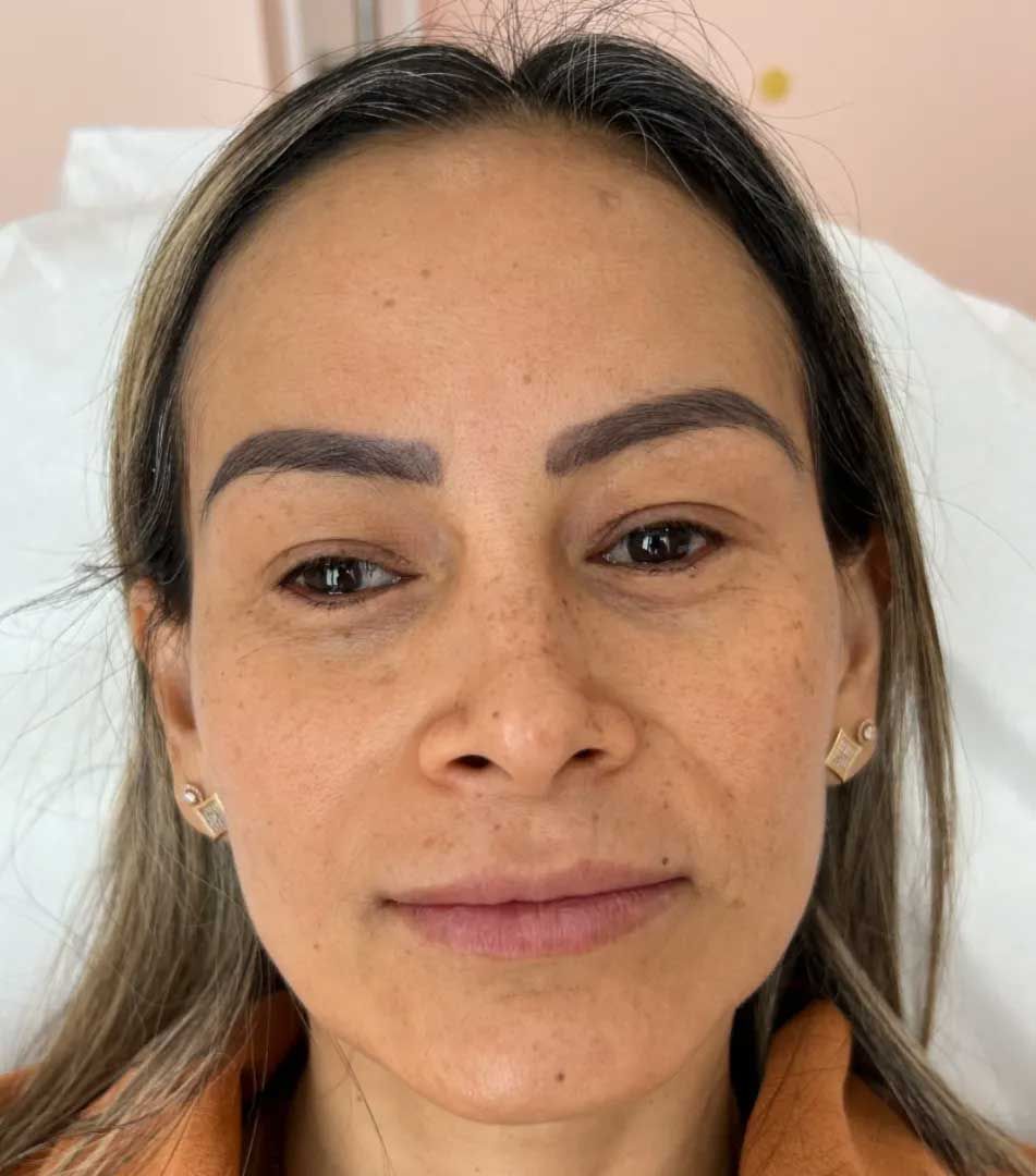 Woman with brown hair and eyebrows, wearing a peach shirt, looking forward. Light skin, slight smile.