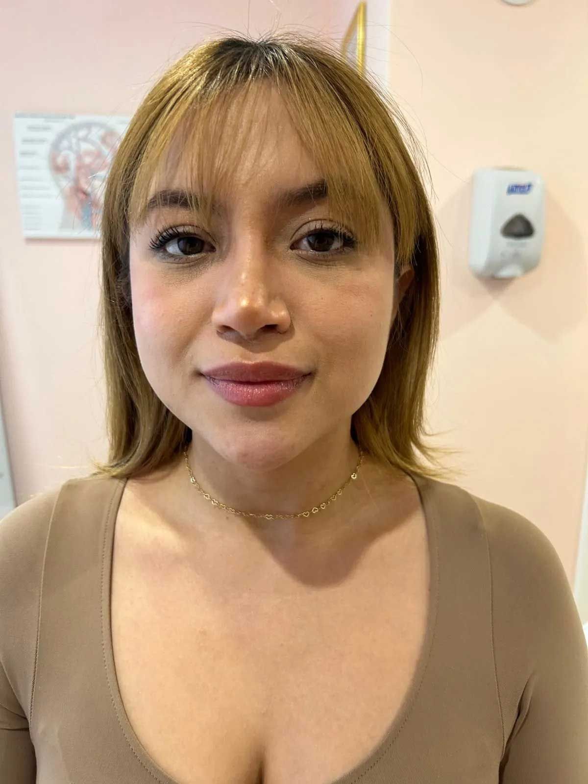 Woman with bangs, wearing a beige top and gold necklace, looking at the camera in a pink room.