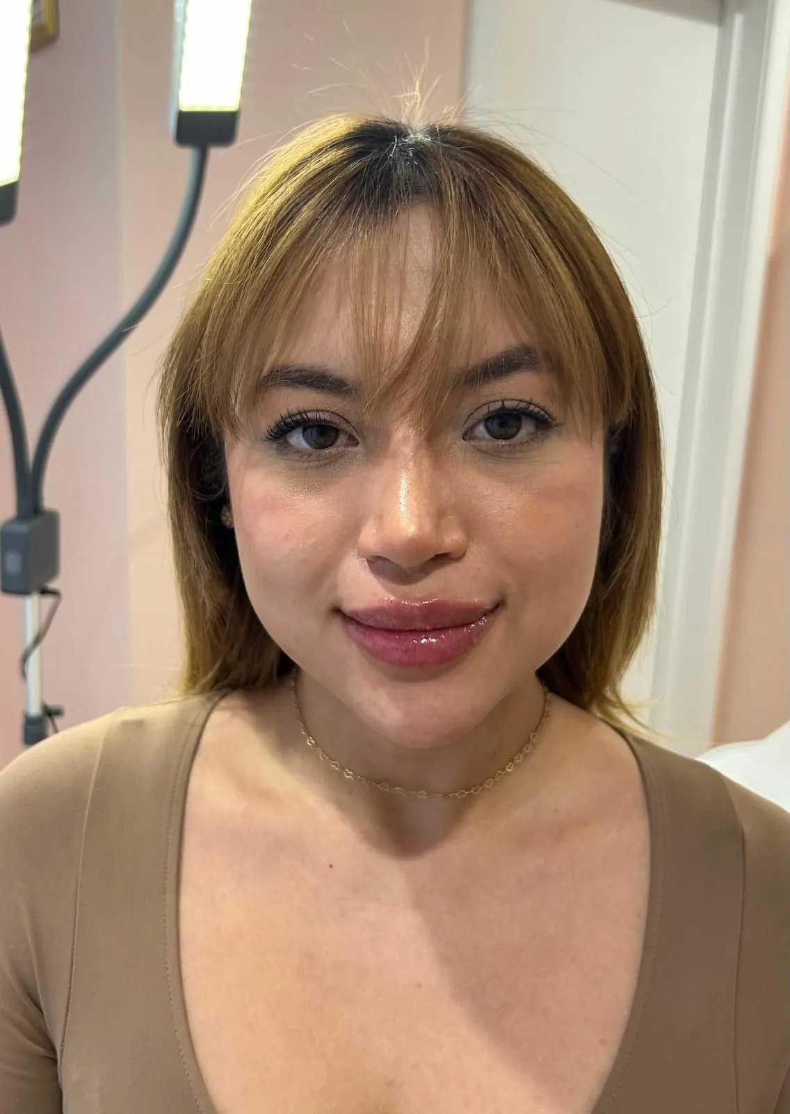 Woman with light brown hair and lip gloss, wearing a tan top, smiles, near a bright light.