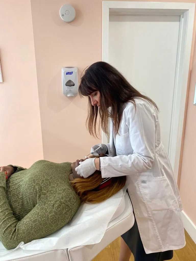 Woman in lab coat performing a procedure on a patient lying on a white table. Pink walls, white door.