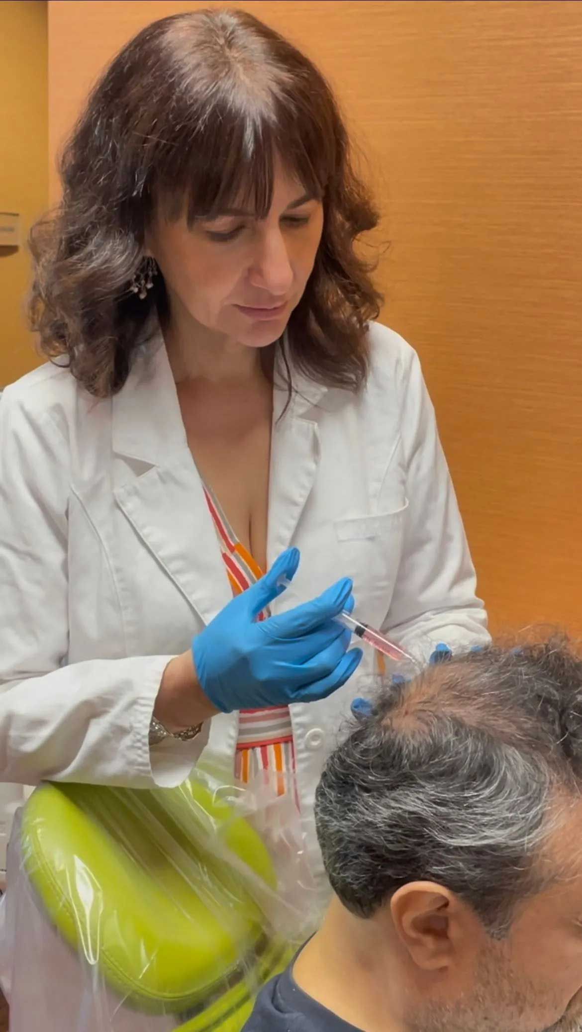 A woman in a white lab coat, wearing gloves, injecting a man's scalp.