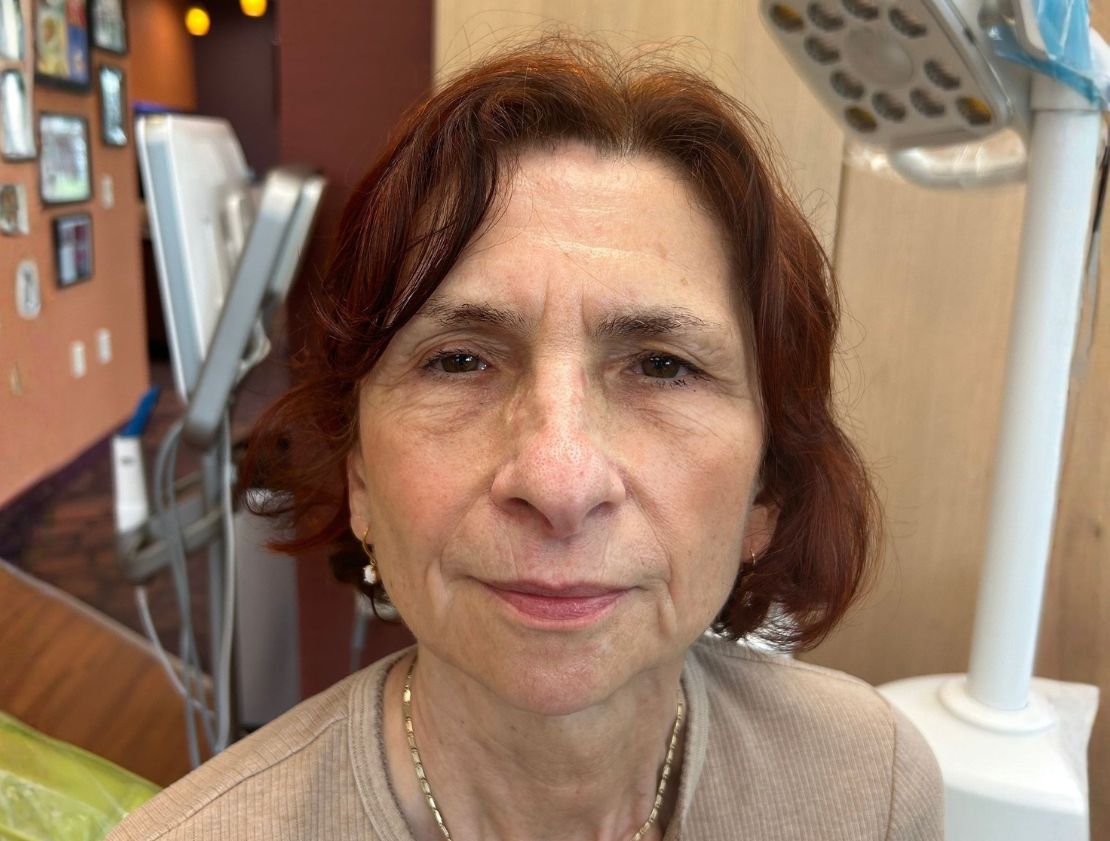 Woman in beige sweater at a dental office, posing for the camera, dental equipment visible.