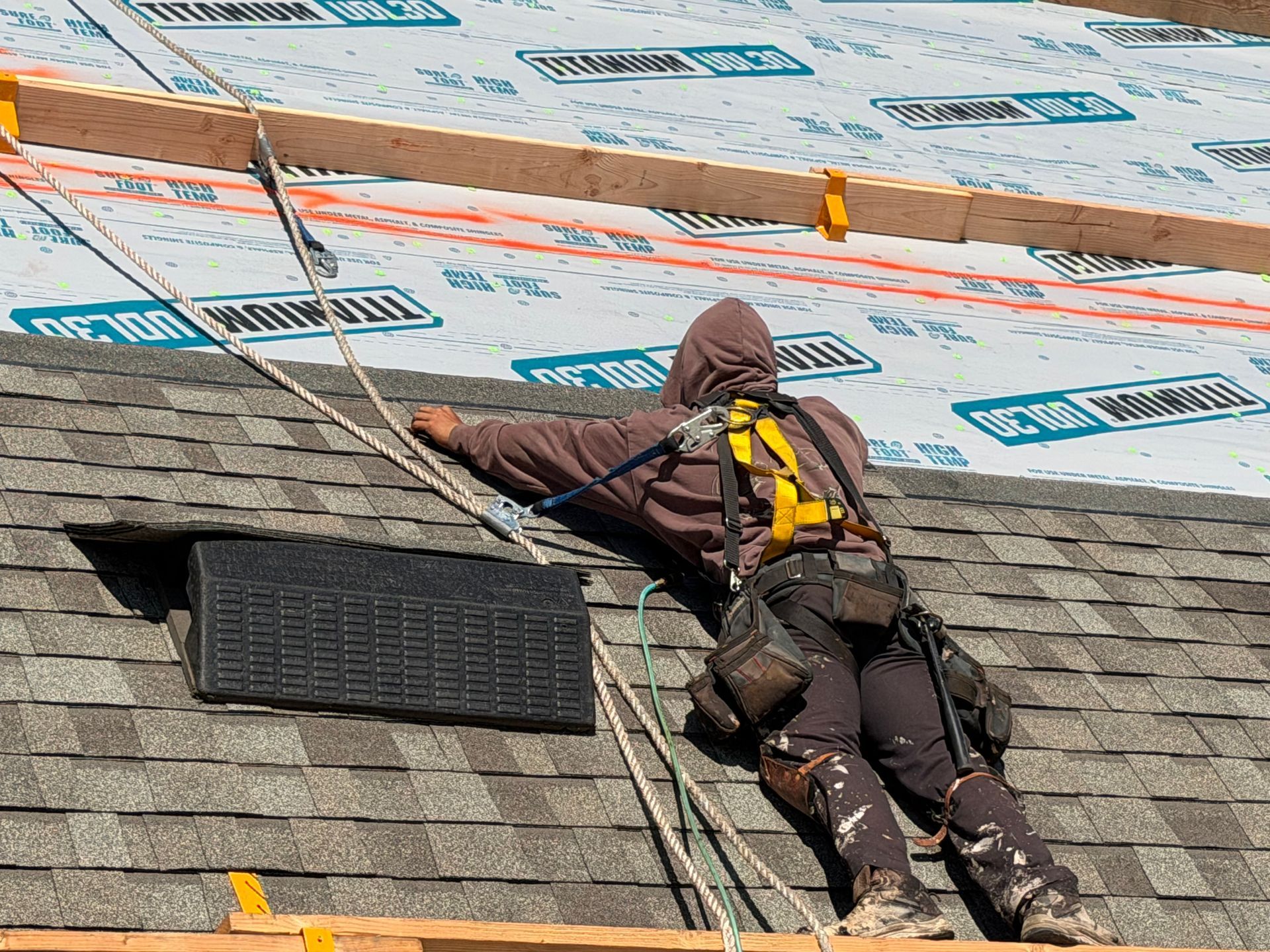 Worker on a shingled roof, secured by ropes, installing or repairing roofing underlayment.